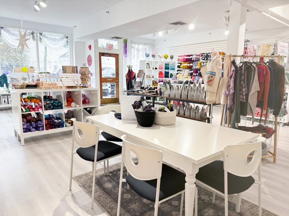 A brightly lit yarn and craft store, home of Circle of Stitches, features shelves of colorful yarn and supplies. Centered is a white table with black chairs, baskets of yarn on top, perfect for Stitch Night gatherings in the natural light by the entrance.
