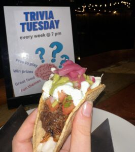 A hand holds a taco with guacamole and onions in front of a Trivia Tuesday sign, as string lights shine in the dark—perfect for a cozy Sevenings at CCE Nightlife in Essex County.