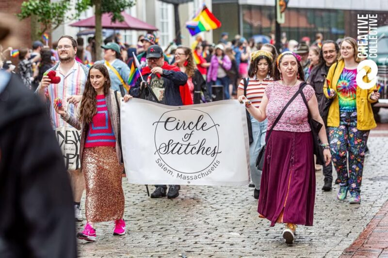 A group walks in a parade holding a “Circle of Stitches Salem, Massachusetts” banner. Many wear colorful clothing and rainbow accessories, celebrating pride. Online quilting and sewing classes unite the community along the cobblestone street.