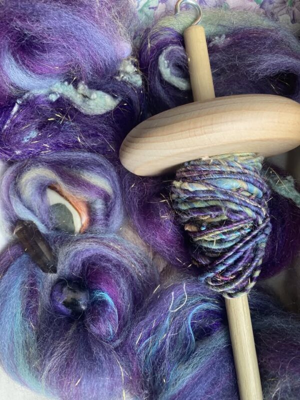 A wooden drop spindle with spun multicolored yarn sits on top of unspun wool roving in shades of purple, blue, and teal, with gold sparkles visible throughout the fibers.
