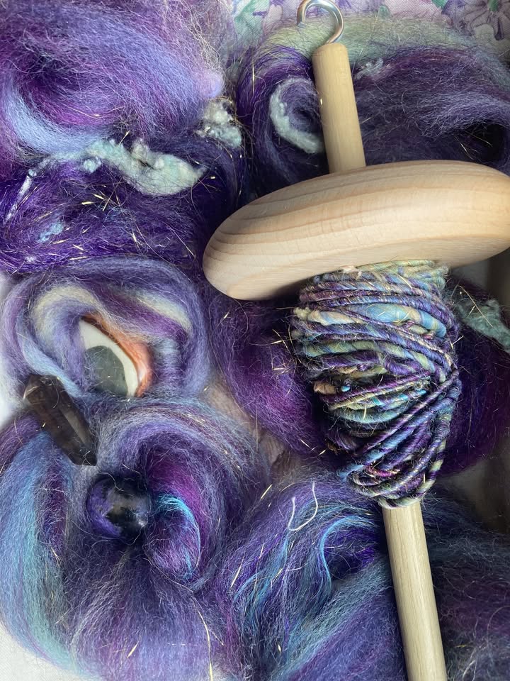 A wooden drop spindle with spun multicolored yarn sits on top of unspun wool roving in shades of purple, blue, and teal, with gold sparkles visible throughout the fibers.