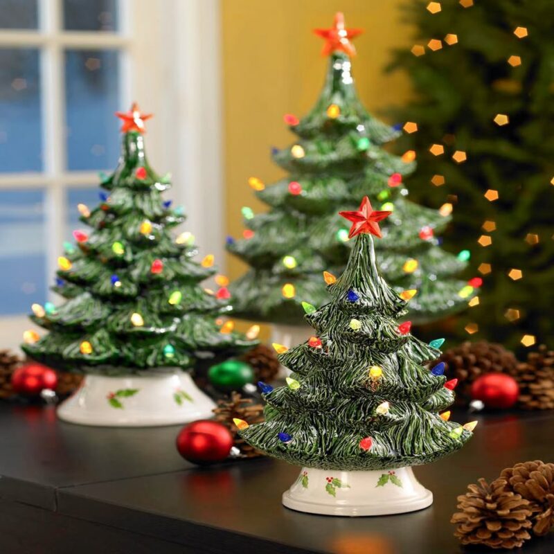 Three ceramic Christmas trees with colored lights and red star toppers from Hestia Designs are displayed on a dark table. Pinecones and red ornaments surround them, with a blurred green tree and window in the background.