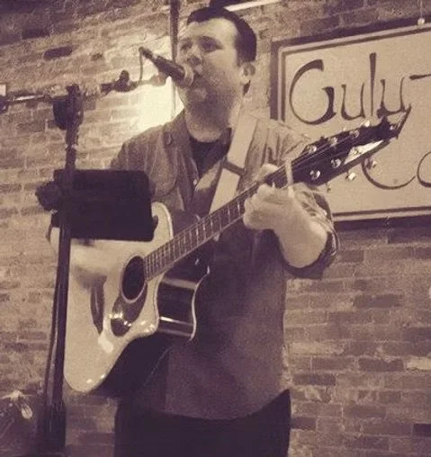 Kevin Farren performs live music, singing and playing acoustic guitar into a microphone indoors. A brick wall and part of a “Guly” sign appear behind him in this sepia-toned image. No cover is required for this intimate performance.