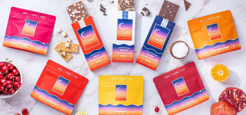 Assorted Coast Cannabis Co. edibles in red, orange, and yellow packaging and chocolate bars are arranged on a white surface, surrounded by fruit, coconut, graham crackers, chocolate chunks, and cherries for a vibrant Coast Cannabis experience.