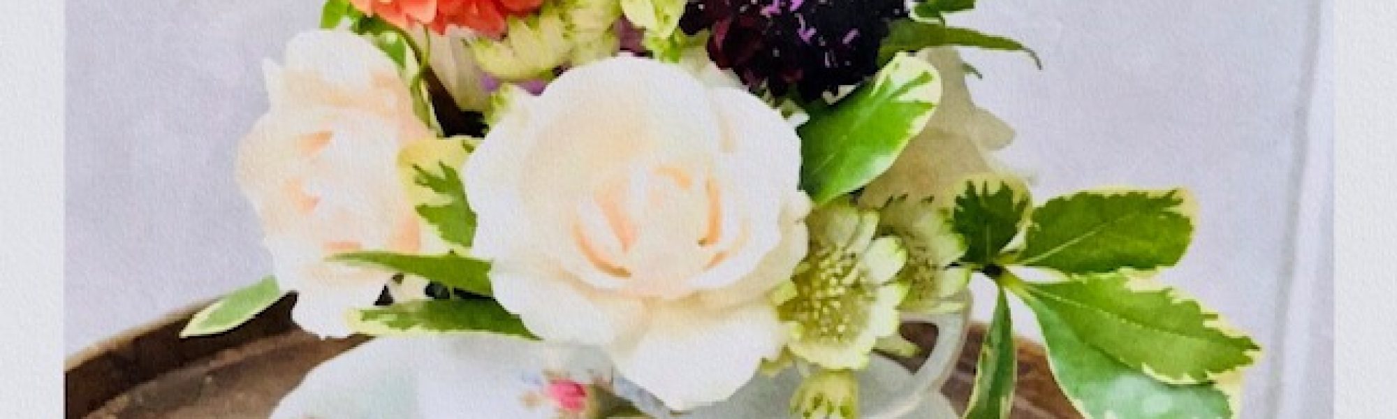 A charming floral design: a small bouquet of white, pink, red, and dark purple flowers with green leaves arranged in a floral teacup, placed on a round tray against a neutral background—perfect inspiration for any workshop.