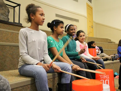 Four girls, ages 8-12 yrs, sit on carpeted steps at a Youth Club drop-in program, holding drumsticks and plastic buckets as they engage in a collaborative drumming activity. More children are visible in the background.