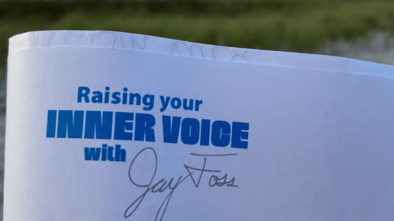 A close-up of a white paper document features “Raising your INNER VOICE with” in blue, the handwritten signature “Jay Foss,” and faint pencil writing. In the soft-focus green background, the scene highlights Jay Foss Music’s creative spirit.