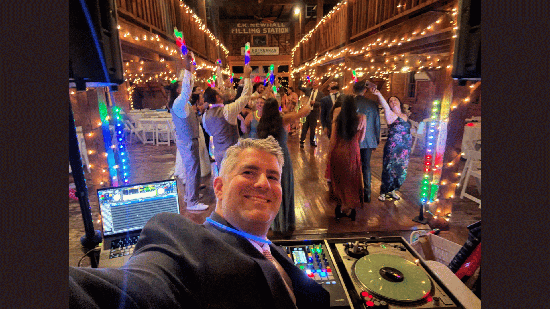 A DJ takes a selfie in front of his turntable at a wedding reception, guests dancing under string lights in a rustic venue. The DJ booth displays equipment and a laptop with music software, reminiscent of the energy at Meet the Member: Salem Horror Fest.