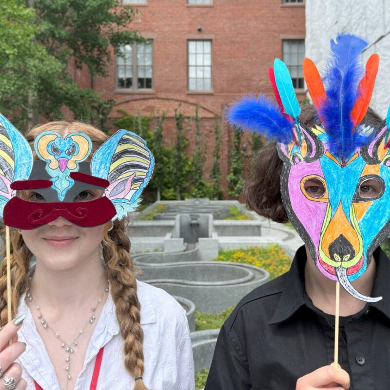 Two people stand side by side outdoors, each holding a brightly decorated animal mask in front of their face. One mask has mouse-like features, while the other is colorful with feathers. A brick building and greenery appear in the background.