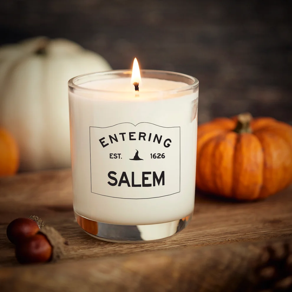 A lit WICKCRAFT white candle in a glass container labeled “Entering Salem Est. 1626” sits on a wooden surface with small pumpkins and chestnuts nearby, ready to inspire your next pour your own Halloween candle creation.