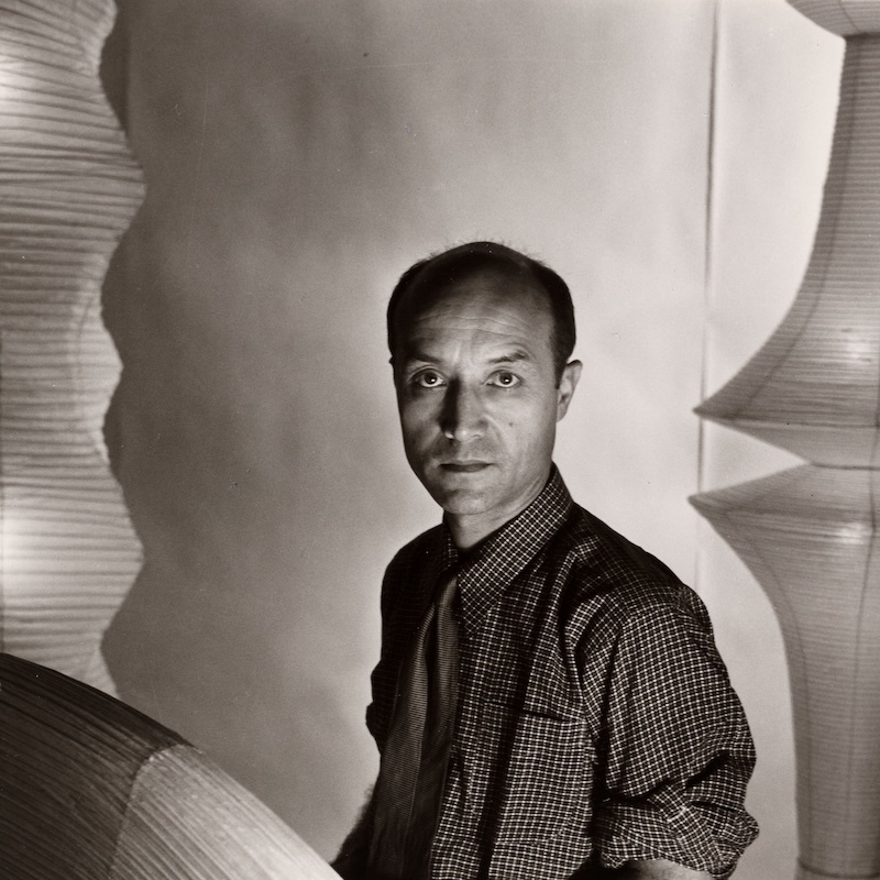 A man with a receding hairline, wearing a checkered shirt and tie, stands indoors among large, sculptural, paper-like lamps. The photo is in black and white with dramatic lighting and shadows. The man looks directly at the camera.