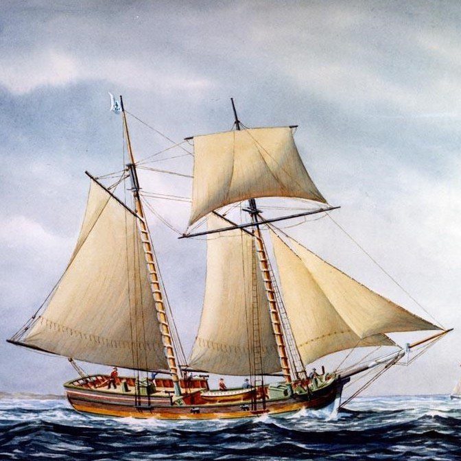 A two-masted sailing ship with tan sails moves across the sea under a cloudy sky. Several people are visible on the deck, and a small flag flies at the top of the front mast.