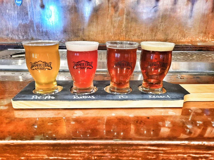 A wooden flight board holds four small glasses of different colored beers, labeled Pit-Pa, Rubus, PO, and Kesell, in front of a metallic bar background. The glasses display the Granite Coast Brewing logo.