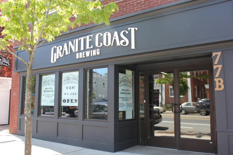 Exterior of Granite Coast Brewing with large windows, a brick facade, and a sign reading “YES! WE ARE OPEN.” A tree is on the sidewalk, and reflections of parked cars and neighboring buildings are visible in the glass.