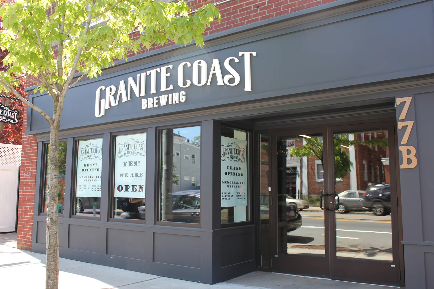 Exterior of Granite Coast Brewing with large windows, a brick facade, and a sign reading “YES! WE ARE OPEN.” A tree is on the sidewalk, and reflections of parked cars and neighboring buildings are visible in the glass.