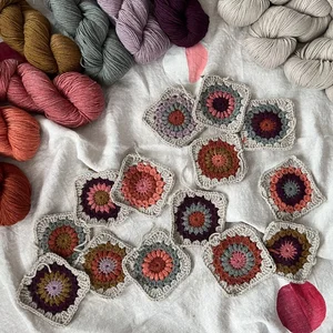 Several colorful crochet granny squares are laid out on a white fabric surface, surrounded by yarn in pink, purple, gold, gray, and cream—perfect inspiration if you want to learn to crochet or need ideas for your next project. Pink flower petals are scattered nearby.