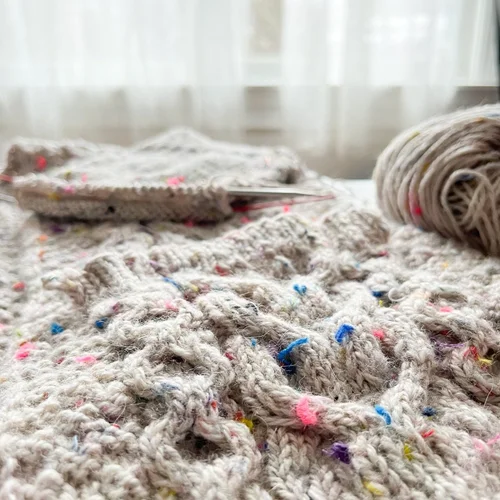 Close-up of a knitted fabric with a speckled pattern in pastel colors. A knitting needle is visible, and a ball of yarn sits nearby. The softly lit background sets the scene—perfect for those ready to learn to crochet or start a new crochet project.