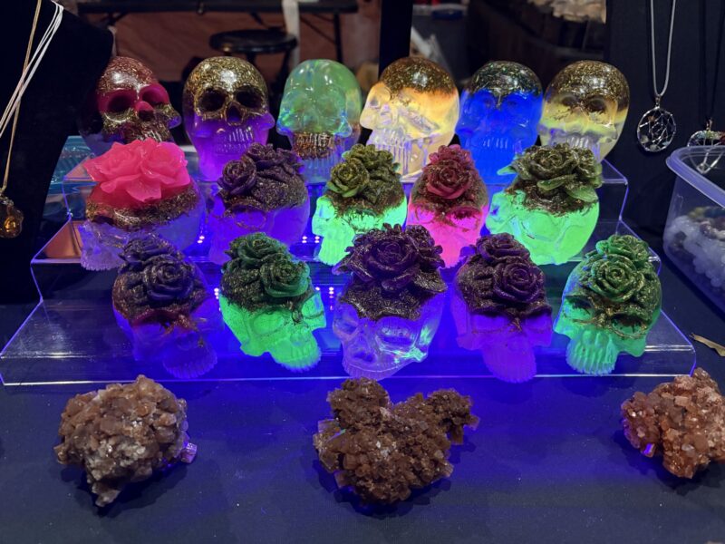 A Positively Horrific display of glass skulls with colorful LED lights and large roses on top, arranged on a clear stand. Brown crystal clusters and various jewelry items complement the eerie yet vibrant table setting.
