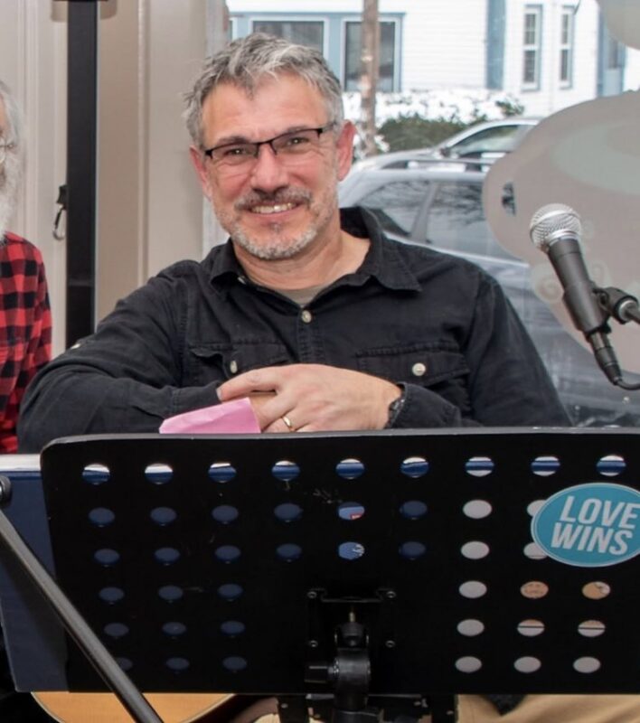 A man with gray hair and glasses sits behind a music stand with a “LOVE WINS” sticker. He is smiling, wearing a black shirt, and holding a pink item. A microphone is positioned in front of him and a guitar is partially visible. A window and car are in the background.