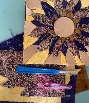 A quilt block with a sunburst pattern in yellow, gold, and brown fabrics lies next to another brown and yellow patch. A blue seam ripper and a pink plastic sewing clip are also visible on the surface.