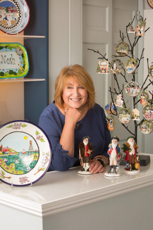 A woman with light brown hair and a blue top stands behind a counter displaying Hestia Creations’ festive figurines and decorative plates. A small tree holds hand-painted ornaments, adding to the celebration of their 40th Anniversary.