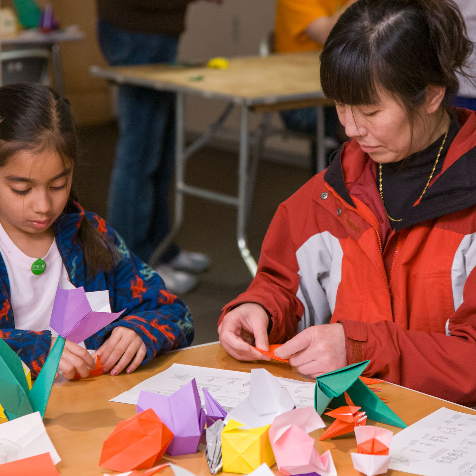 An adult and a child sit at a table folding colorful origami paper. Several finished origami models and instruction sheets are on the table. Both people are focused on their paper crafts. The background shows another table and a person standing.