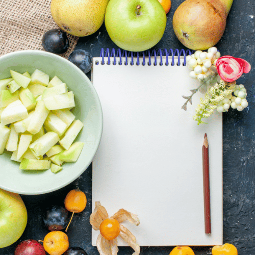 A blank notepad with a pencil beside a bowl of chopped green apples, perfect for jotting notes during your free nutrition class, surrounded by fresh fruits and a small bouquet of flowers on a dark surface.