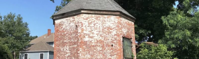 A small, round brick building with a weathered exterior and gray shingled roof stands in a grassy area, inviting visitors to explore its history during Powder House Tours as part of the Trails & Sails event, with trees and a house in the background.