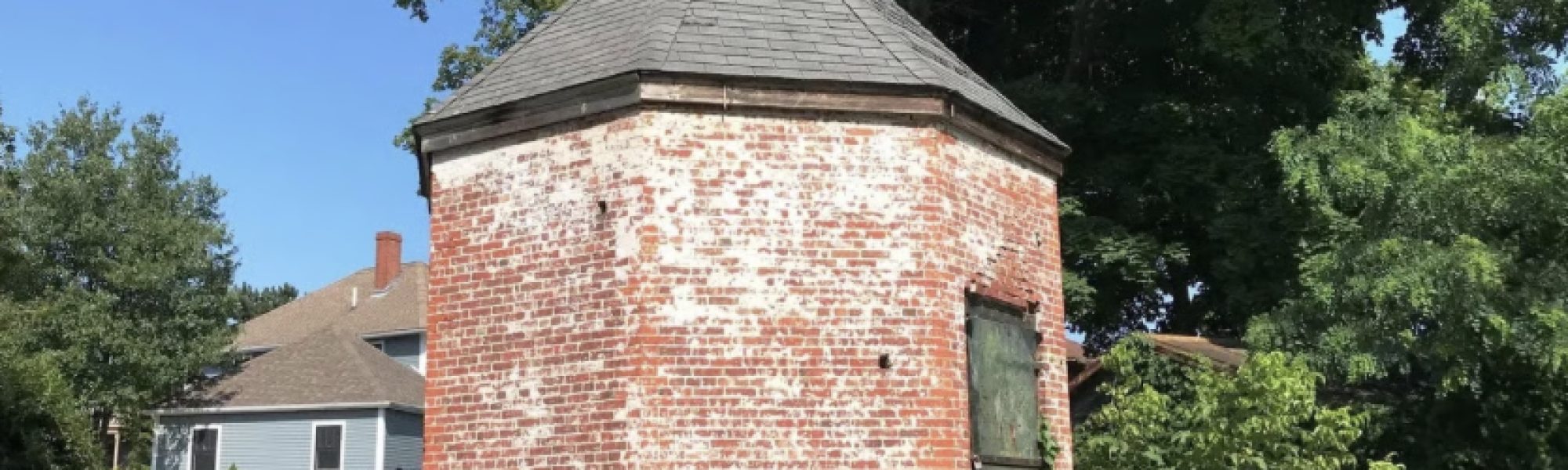 A small, round brick building with a weathered exterior and gray shingled roof stands in a grassy area, inviting visitors to explore its history during Powder House Tours as part of the Trails & Sails event, with trees and a house in the background.
