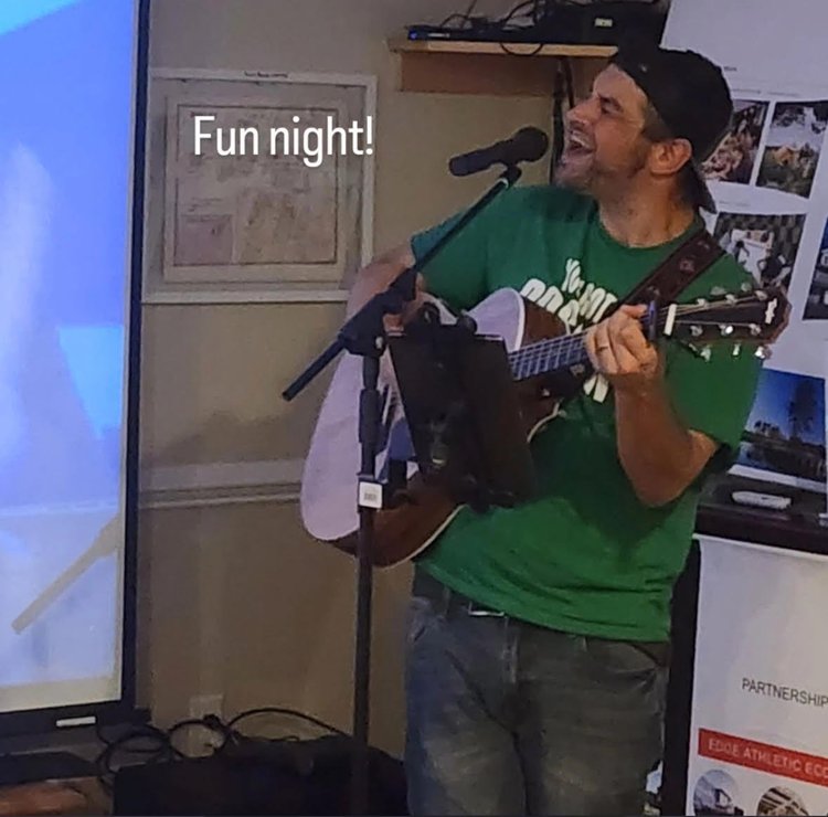 A man wearing a green shirt and black cap sings into a microphone while playing an acoustic guitar indoors. There are posters and a projector screen in the background. The text “Fun night!” appears at the top left.