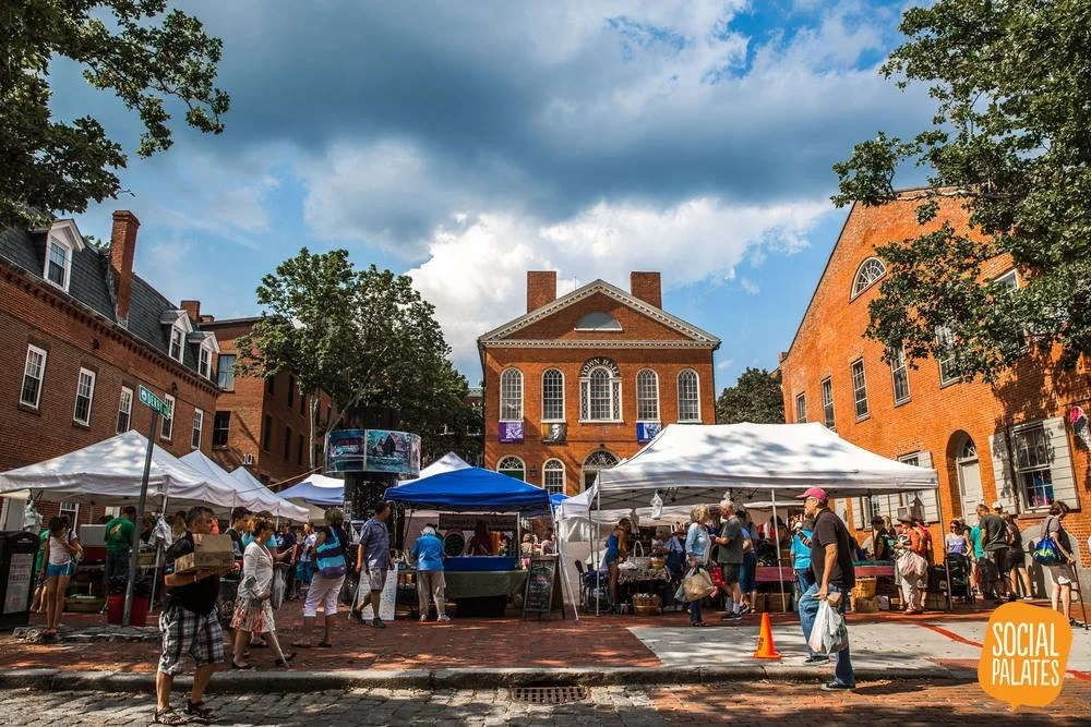 A farmers market with white tents and people browsing stands is set up on a cobblestone street in front of a red brick building under a partly cloudy sky. Trees frame the scene, and a “SOCIAL PALATES” logo appears in the lower right corner.