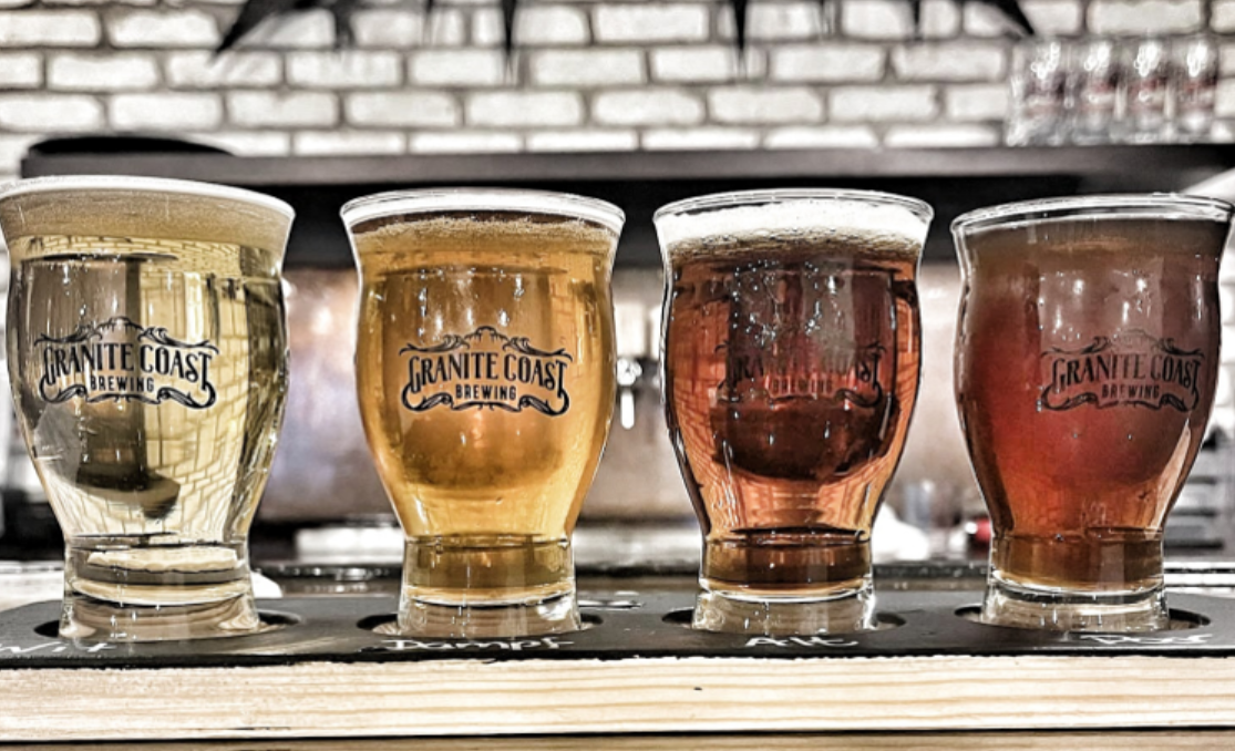 Four Granite Coast Brewing beers, each a unique style and color, are arranged in a row for a guided tasting. The wooden tray sits before a blurred brick wall and shelves, inviting you to explore every flavor note.