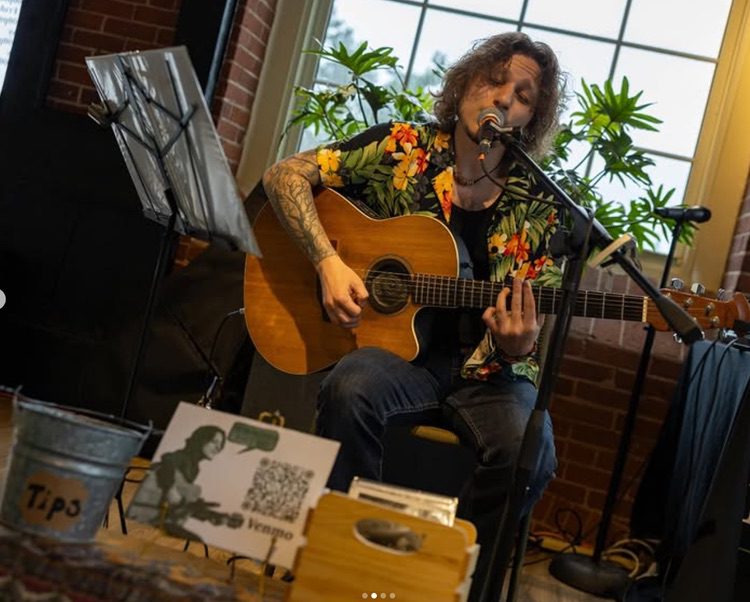 A person with curly hair plays an acoustic guitar and sings into a microphone indoors. They wear a floral shirt. A music stand, tip jar, and a sign with a QR code are visible in the foreground. Green plants and a window are in the background.