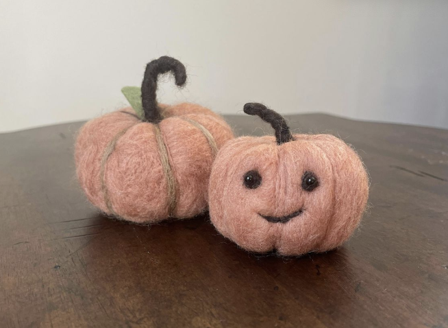 Two small, peach-colored felt pumpkins sit on a wooden surface at the Hutch. One pumpkin has a smiling face, and both have brown stems. A small green leaf is attached to the pumpkin without a face; the background is plain and out of focus.