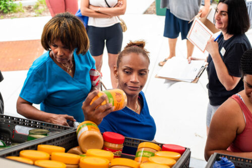 Several people stand around bins of peanut butter jars at a Pop-Up event in Cook Street Park. A woman in blue scrubs inspects a jar while others wait nearby, holding papers or clipboards—likely at a food distribution or pantry event.