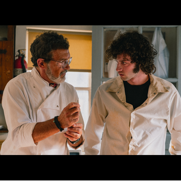 Two men in white coats stand in a kitchen or lab setting, engaged in conversation. The older man gestures with his hand while speaking, and the younger man listens intently. Shelves and a window are visible in the background.