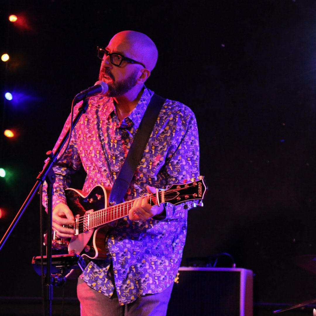 A man with glasses and a patterned shirt plays an electric guitar and sings into a microphone on stage. Colored lights are visible in the background, and an amplifier sits behind him. The stage is dimly lit.