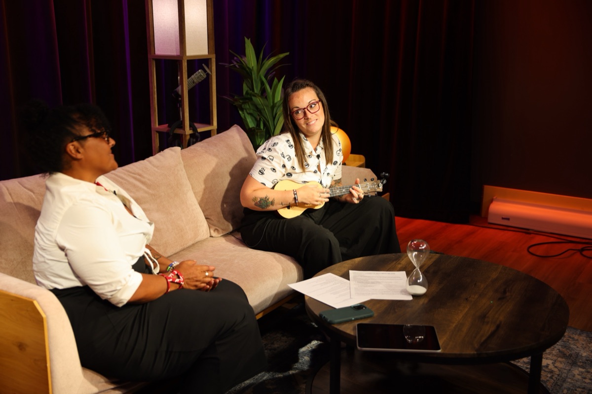 Amanda "A" Larson plays a ukulele on a couch while Mikki Wilson laughs from the other end, notes and an hourglass on the coffee table.