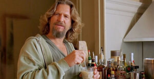 A man with long hair and a beard stands in a kitchen, holding a drink and a stirring stick. Wearing a light robe over a T-shirt, he’s ready for Essex County nightlife, with several bottles of alcohol arranged on the counter beside him.