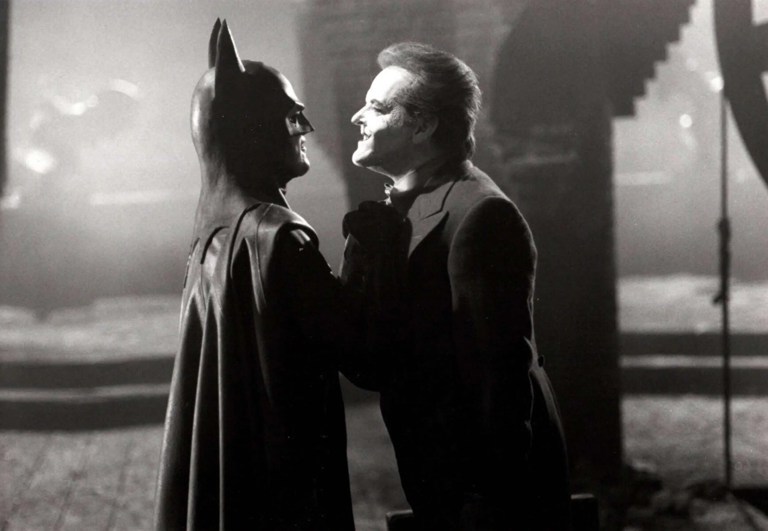 A person dressed as Batman grabs another person dressed as the Joker by the collar in a dramatic black and white scene, with gothic architecture visible in the background.