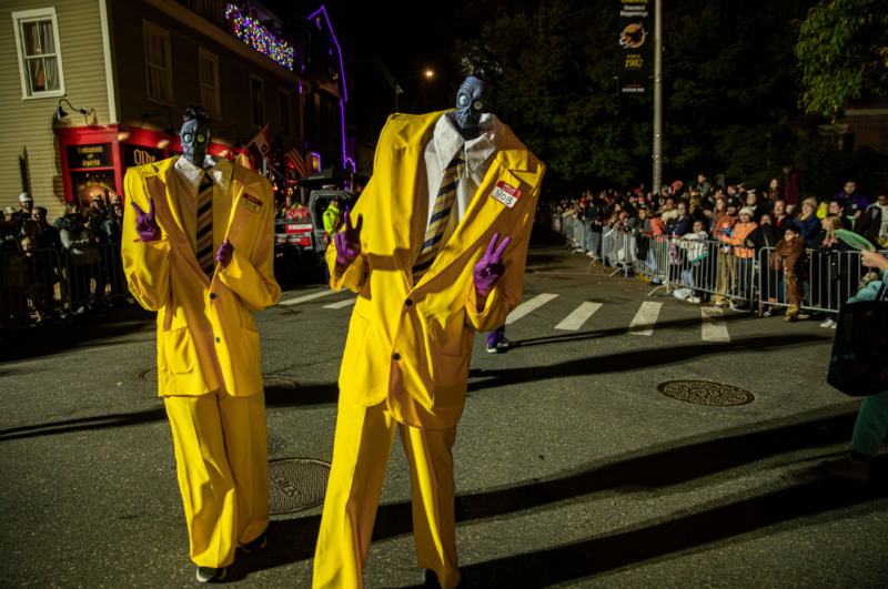 Two people in oversized yellow suits, black masks with bulging eyes, and purple gloves pose with peace signs on a street at night during the Haunted Happenings Grand Parade, as a crowd watches from behind barriers.