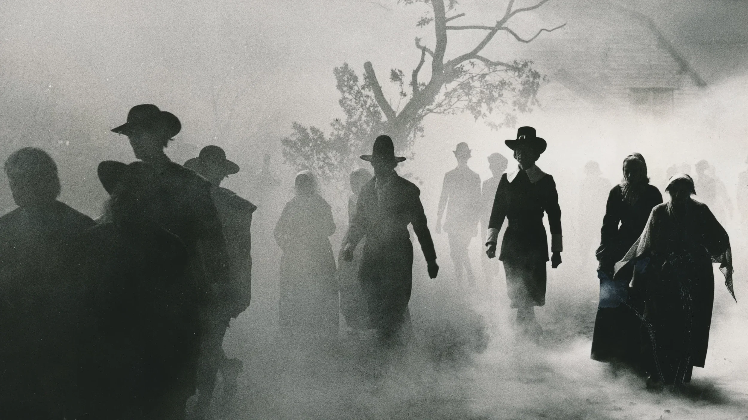 A group of people in old-fashioned clothing and wide-brimmed hats walk through thick fog. Silhouetted figures and a bare tree are in the background, creating a mysterious, eerie atmosphere.