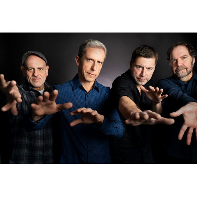 Four middle-aged men stand in a row against a dark background, extending their arms and hands toward the camera. They wear casual shirts and serious expressions. The image is well-lit, with focused lighting on their faces and upper bodies.