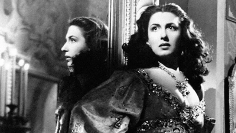 A woman in an ornate dress stands beside a mirror, her reflection visible. She has long, dark hair and wears a necklace. The background appears to be a decorated, elegant room. The photo is in black and white.