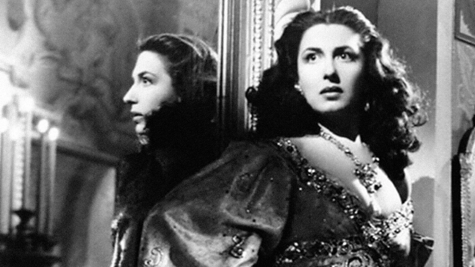 A woman in an ornate dress stands beside a mirror, her reflection visible. She has long, dark hair and wears a necklace. The background appears to be a decorated, elegant room. The photo is in black and white.