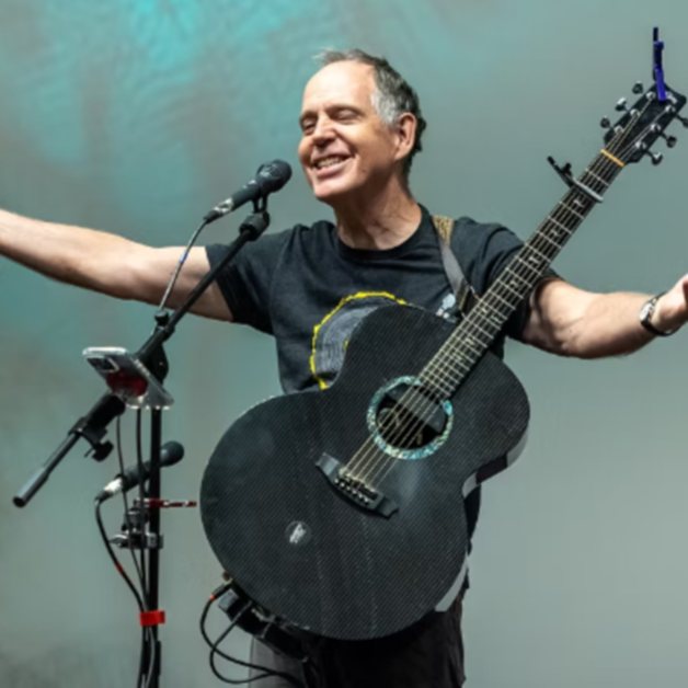 David Wilcox stands on stage with a black acoustic guitar, arms outstretched and smiling. He wears a black T-shirt and is in front of a microphone setup. The background is plain and softly lit.