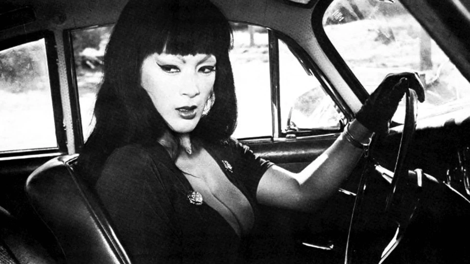 A woman with straight dark hair and bangs sits in the driver’s seat of a car, wearing a low-cut dress and gloves. She looks to her left with a serious expression. The image is in black and white.
