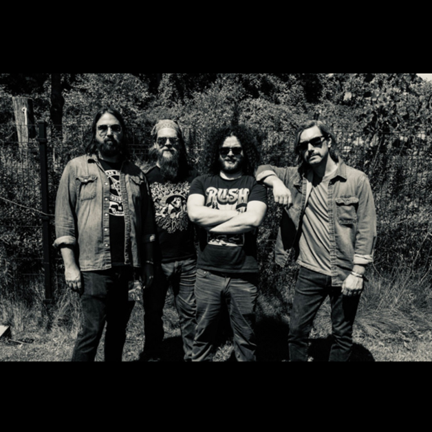 Four men, including Nate Wilson from Ghosts of Jupiter, stand outdoors in front of a fence and trees. Three have beards; one wears sunglasses. They sport casual jackets and graphic tees in this black-and-white image.