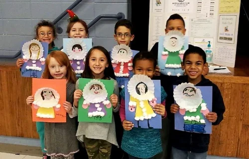 A group of children stands indoors at the Kids Club, each holding up a colorful craft project with a cotton-ball collar. Smiling, they pose in front of a stage area with posters behind them—perfect for fun Kids Activities and program registration events.