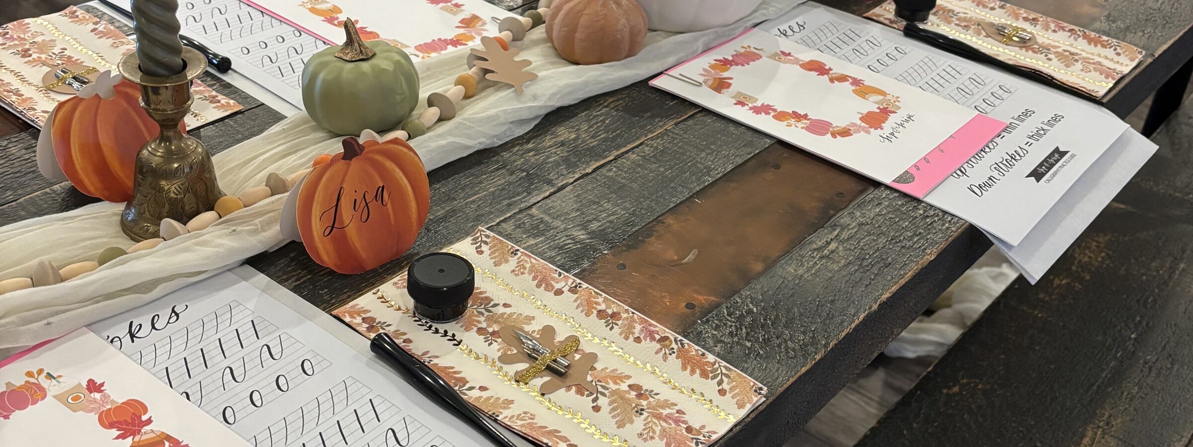 A wooden table set for a fall-themed calligraphy class, featuring small decorative pumpkins, worksheets with practice alphabets, an ink pot, a pen, candles, and autumn leaf placemats. Each place setting includes a name card and a worksheet.